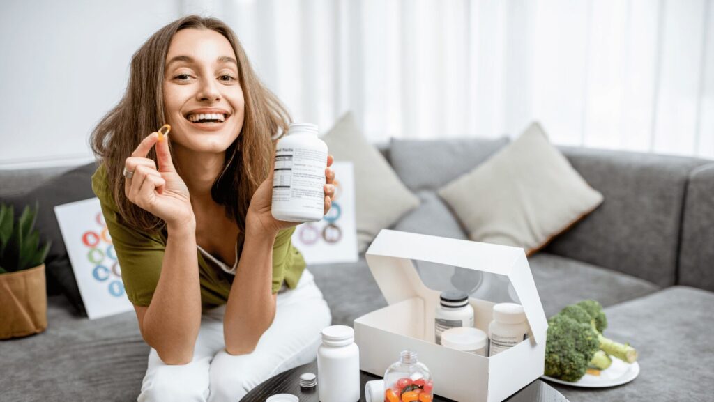 Lachende Frau hält eine Vitamin-D-Kapsel und eine Flasche Nahrungsergänzungsmittel in der Hand, symbolisiert die Bedeutung von Vitamin D zur Vorbeugung von Vitamin-D-Mangel-Symptomen.