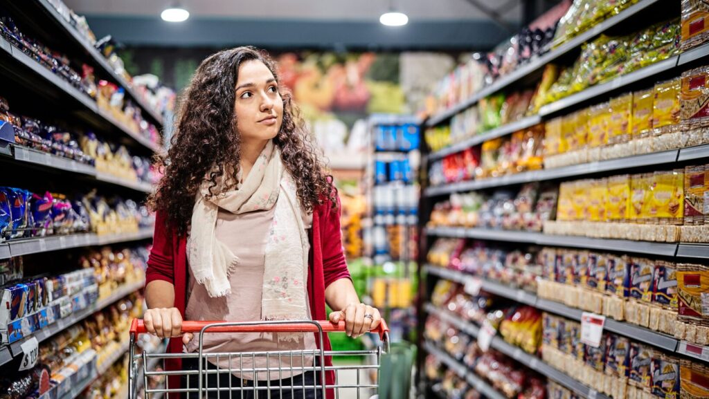 Frau mit Einkaufswagen im Supermarkt, umgeben von Regalen voller verarbeiteter Lebensmittel.