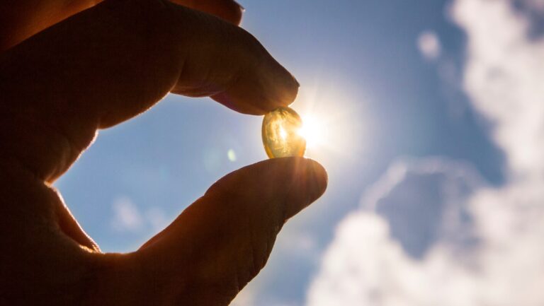 Hand hält eine Vitamin-D-Kapsel vor die Sonne, symbolisiert die Wichtigkeit von Sonnenlicht zur Vorbeugung von Vitamin-D-Mangel-Symptomen.