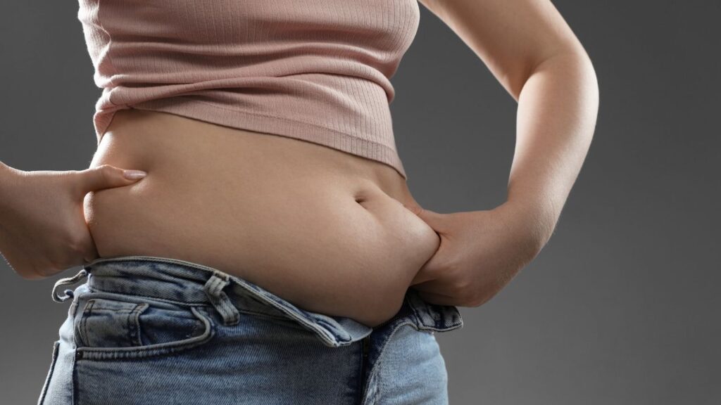 Frau mit geöffnetem Jeansbund hält sichtbares Fett am Bauch – typisches Beispiel für subkutanes Fett im Bauchbereich